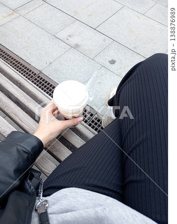 A girl is holding a cup of coffee in her hands, sitting on a bench. Relaxing moment outdoors with warm drink and peaceful atmosphere A girl is holding a cup of coffee in her hands, sitting on a bench. Relaxing moment outdoors with warm drink and peaceful atmosphere 138876989