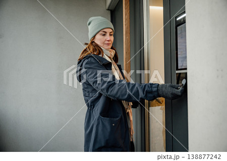 Woman using a doorbell at an entrance of a modern building on a chilly day 138877242