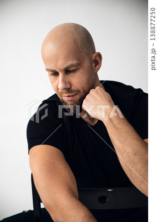 Man with a bald head and beard sits thoughtfully in a studio setting Man with a bald head and beard sits thoughtfully in a studio setting 138877250