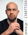 Person with shaved head sits with hands clasped in front of a plain background indoors 138877268