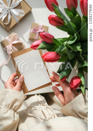 Vertical lifestyle mockup of woman holding blank white card with craft envelope over table with red tulips and craft gifts on white table under bright natural sunlight 138877690