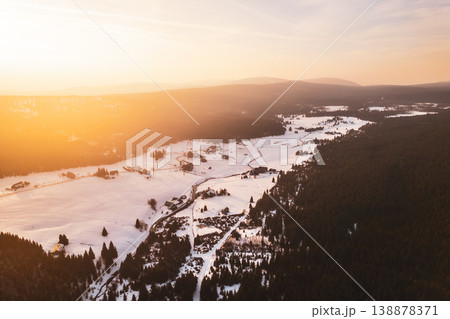 The scene shows Jizerka mountain village covered in snow during winter. A drone captures the village layout, the trees, and the snow-covered land under a sunset sky. The scene shows Jizerka mountain village covered in snow during winter. A drone captures the village layout, the trees, and the snow-covered land under a sunset sky. 138878371