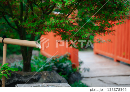 Traditional bamboo fountain in a lush Japanese garden featuring maple leaves and orange fence Traditional bamboo fountain in a lush Japanese garden featuring maple leaves and orange fence 138878563