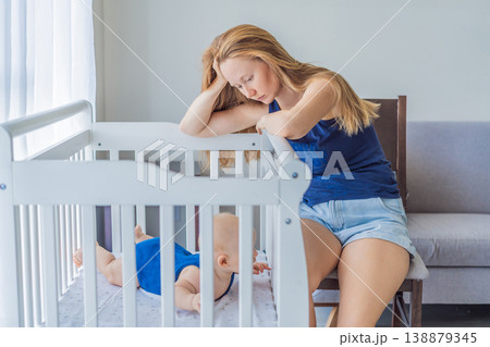 Very tired mother sitting by baby crib at home feeling exhausted. Sleep deprived mom caring for newborn at night. Parenting fatigue, motherhood stress, childcare and real family life concept 138879345