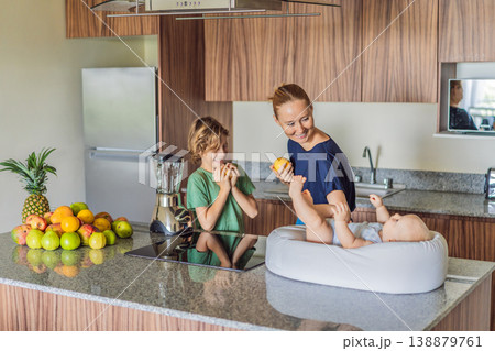 Mother and son preparing smoothie in kitchen while baby lies in cozy lounger on table watching. Family cooking together, healthy lifestyle, parenting, sibling bonding and everyday life concept Mother and son preparing smoothie in kitchen while baby lies in cozy lounger on table watching. Family cooking together, healthy lifestyle, parenting, sibling bonding and everyday life concept 138879761
