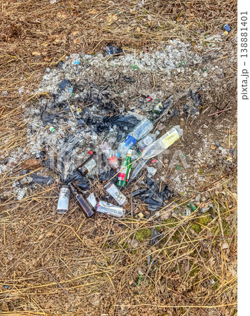 A messy heap of trash, including glass bottles, plastic, and burnt debris, pollutes the ground in a forest. Concept of environmental damage.. High quality photo A messy heap of trash, including glass bottles, plastic, and burnt debris, pollutes the ground in a forest. Concept of environmental damage.. High quality photo 138881401