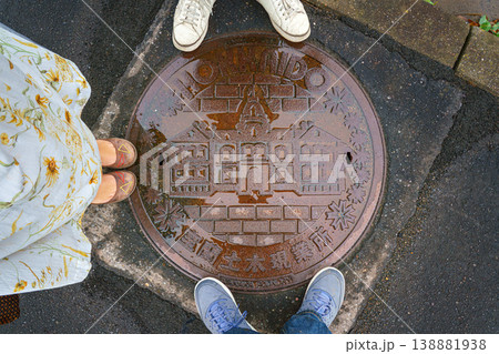 Muroran, Hokkaido, Japan - Oct 4 2024, close up view of a manhole cover with an image of a building and the inscription Hokkaido, with three people's feet standing around, Muroran, Japan Muroran, Hokkaido, Japan - Oct 4 2024, close up view of a manhole cover with an image of a building and the inscription Hokkaido, with three people's feet standing around, Muroran, Japan 138881938