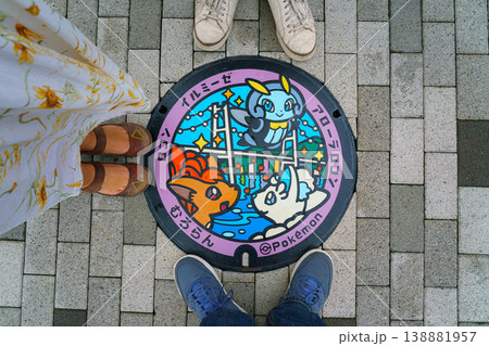 Muroran, Hokkaido, Japan - Oct 4 2024, A close-up view of a manhole with a color image of three cute Pokemon on background of Hakucho Bridge, with three people's feet standing around, Muroran, Japan Muroran, Hokkaido, Japan - Oct 4 2024, A close-up view of a manhole with a color image of three cute Pokemon on background of Hakucho Bridge, with three people's feet standing around, Muroran, Japan 138881957
