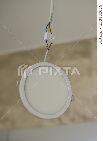 A modern round white LED recessed panel light hanging by exposed electrical wires and a terminal block from a ceiling during installation. High quality photo A modern round white LED recessed panel light hanging by exposed electrical wires and a terminal block from a ceiling during installation. High quality photo 138882056