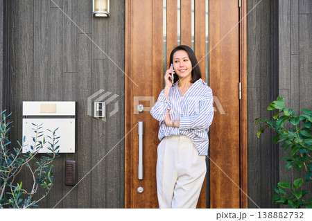 一軒家の玄関前でスマホで通話しながら笑顔を見せる外出前の日本人女性 一軒家の玄関前でスマホで通話しながら笑顔を見せる外出前の日本人女性 138882732