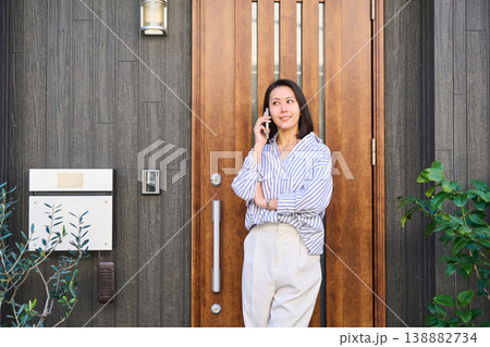 一軒家の玄関前でスマホで通話しながら笑顔を見せる外出前の日本人女性 138882734