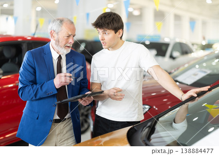 Elderly auto salesman advises a guy, answering his questions and showing detailed information Elderly auto salesman advises a guy, answering his questions and showing detailed information 138884477