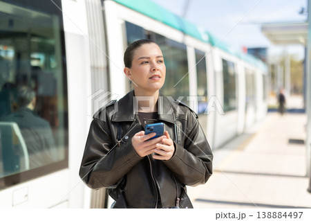 Portrait of girl in black jacket with mobile phone in her hands at tram stop on sunny spring day Portrait of girl in black jacket with mobile phone in her hands at tram stop on sunny spring day 138884497