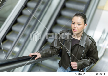 Positive young woman in black leather jacket riding up an escalator at metro station Positive young woman in black leather jacket riding up an escalator at metro station 138884831