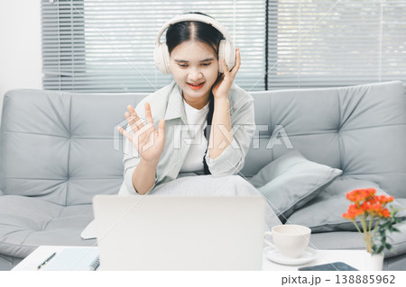 Asian woman wearing headphones and waving during laptop video call at home, showing remote work, online meeting, virtual communication and cozy home office lifestyle in bright living room. Asian woman wearing headphones and waving during laptop video call at home, showing remote work, online meeting, virtual communication and cozy home office lifestyle in bright living room. 138885962