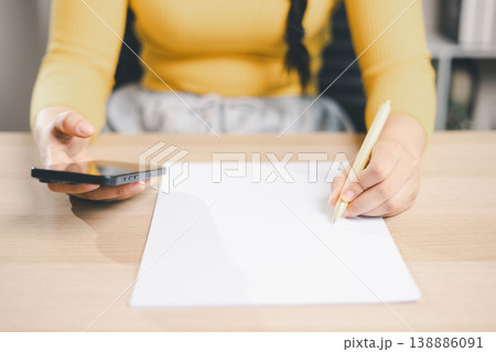 Close up of woman holding smartphone and pen over blank white paper on desk, planning notes or online research. Mobile learning, digital communication, study and workspace concept. 138886091