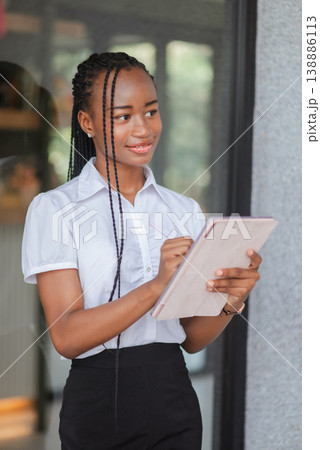 Vertical portrait of a young African woman writing on tablet with stylus while standing outdoors, concept of digital planning, note taking, mobile productivity and modern professional workflow. Vertical portrait of a young African woman writing on tablet with stylus while standing outdoors, concept of digital planning, note taking, mobile productivity and modern professional workflow. 138886113