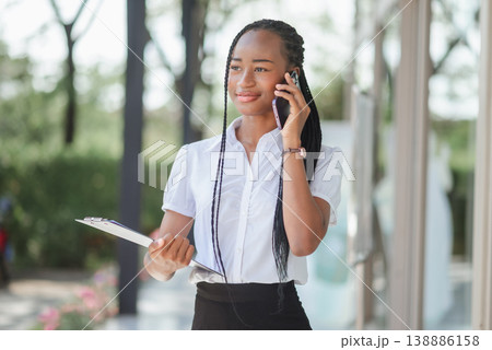 Young African business woman talking on smartphone while holding clipboard outdoors near office, concept of customer contact, professional communication, field work and modern corporate lifestyle. Young African business woman talking on smartphone while holding clipboard outdoors near office, concept of customer contact, professional communication, field work and modern corporate lifestyle. 138886158