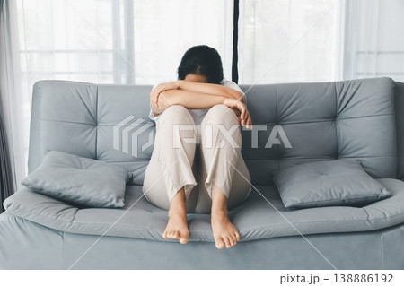 Depressed woman curled up on gray sofa, head down on arms, barefoot at home by bright window. Mental health anxiety, sadness, burnout, loneliness concept, copy space for therapy awareness. depressed 138886192