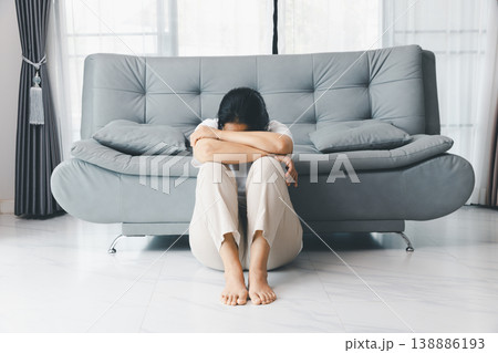 Depressed woman sitting on floor in front of gray sofa, head down on arms and knees, barefoot at home in bright living room. Mental health anxiety, sadness, burnout, loneliness concept. depression 138886193