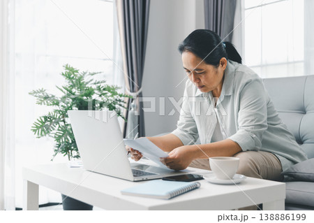 Woman working from home on laptop at coffee table, reading report papers with coffee cup and notebook in bright living room. Remote work, telecommuting, home office productivity concept. remote work Woman working from home on laptop at coffee table, reading report papers with coffee cup and notebook in bright living room. Remote work, telecommuting, home office productivity concept. remote work 138886199