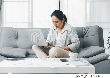 Focused woman working from home on laptop on the sofa, with reports, smartphone and coffee on table in bright living room. Remote work and home office productivity concept for telecommuting. telework 138886200