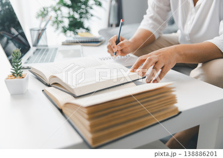 Hands studying at home, reading open textbooks while taking notes beside laptop on bright white desk with stationery and plant. Education, online learning, homework and exam prep concept. learning 138886201