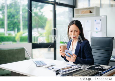 Confident Asian businesswoman using smartphone and drinking coffee at office desk with laptop and documents. Confident Asian businesswoman using smartphone and drinking coffee at office desk with laptop and documents. 138887858