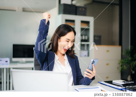 Asian businesswoman celebrates great news on smartphone, smiling with raised fist at office desk. Asian businesswoman celebrates great news on smartphone, smiling with raised fist at office desk. 138887862
