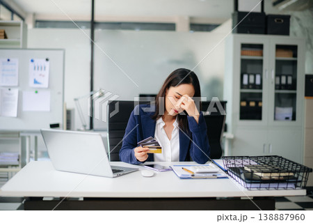 Stressed Asian businesswoman holding multiple credit cards in office, facing financial pressure. 138887960