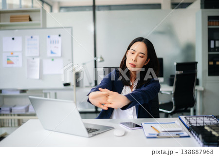 Asian businesswoman stretching arms in office with tired expression, relieving muscle tension from long work hours. Asian businesswoman stretching arms in office with tired expression, relieving muscle tension from long work hours. 138887968