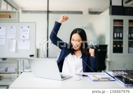 Asian businesswoman celebrating success with fists raised in joy at office desk with laptop. 138887976