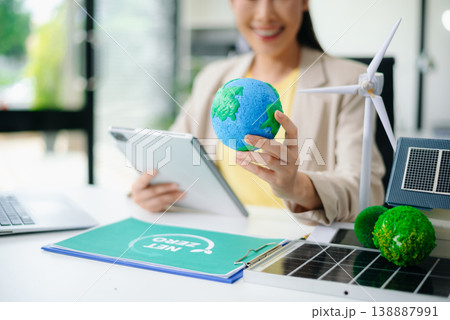Asian woman in office holding earth model beside solar panels and wind turbine. Concept of sustainability, climate change, and clean energy innovation. 138887991
