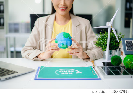 Asian woman in office holding earth model beside solar panels and wind turbine. Concept of sustainability, climate change, and clean energy innovation. 138887992