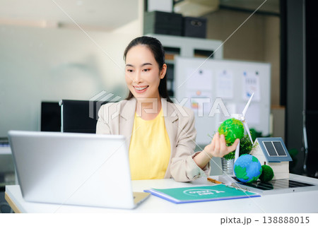 Asian woman working on laptop while holding green earth ball. Surrounded by wind turbine, solar panel, and eco-home models. Concept of clean energy and sustainability. 138888015