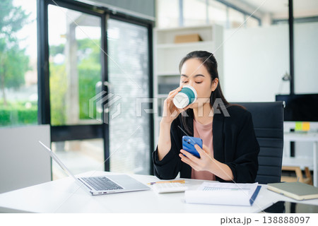 Confident beautiful Asian businesswoman typing laptop computer and digital tablet while holding coffee at office 138888097