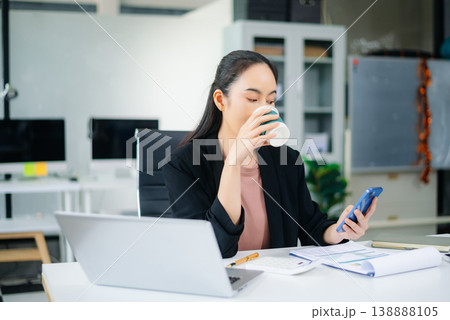 Confident beautiful Asian businesswoman typing laptop computer and digital tablet while holding coffee at office Confident beautiful Asian businesswoman typing laptop computer and digital tablet while holding coffee at office 138888105