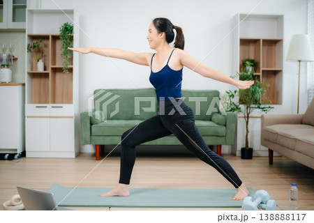 Asian woman practices yoga at home while watching an online tutorial on laptop. Indoor fitness, healthy lifestyle, stretching on mat in cozy modern living room. Asian woman practices yoga at home while watching an online tutorial on laptop. Indoor fitness, healthy lifestyle, stretching on mat in cozy modern living room. 138888117