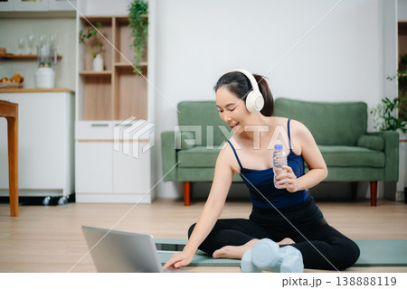 Asian woman practices yoga at home while watching an online tutorial on laptop. Indoor fitness, healthy lifestyle, stretching on mat in cozy modern living room. Asian woman practices yoga at home while watching an online tutorial on laptop. Indoor fitness, healthy lifestyle, stretching on mat in cozy modern living room. 138888119