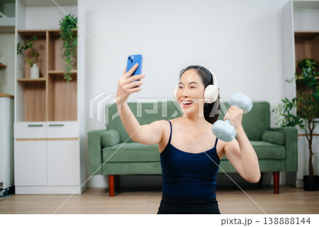 Asian woman practices yoga at home while watching an online tutorial on laptop. Indoor fitness, healthy lifestyle, stretching on mat in cozy modern living room. Asian woman practices yoga at home while watching an online tutorial on laptop. Indoor fitness, healthy lifestyle, stretching on mat in cozy modern living room. 138888144