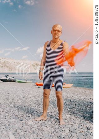 Man swimsuit beach stands on a pebble shore wearing a retro striped bodysuit while waving a vibrant orange fabric during summer vacation Man swimsuit beach stands on a pebble shore wearing a retro striped bodysuit while waving a vibrant orange fabric during summer vacation 138888878