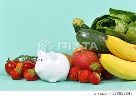 Concept for costs of healthy eating or saving money when buying food. Vegetables and fruits next to piggy bank on blue background with copy space Concept for costs of healthy eating or saving money when buying food. Vegetables and fruits next to piggy bank on blue background with copy space 138889245