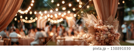 Guests gather under a large wedding tent filled with warm lights. The area is decorated with flowers and creates a festive ambiance for the outdoor celebration at dusk Guests gather under a large wedding tent filled with warm lights. The area is decorated with flowers and creates a festive ambiance for the outdoor celebration at dusk 138891030