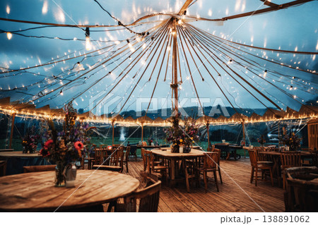 Guests are seated at wooden tables inside a wedding tent. Strings of lights hang from the top. The view shows water and city lights in the background during the evening 138891062