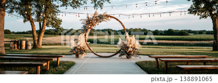 A wedding ceremony is set outdoors with a wooden arch adorned with wildflowers and pampas grass. Chairs are arranged in front of the arch. String lights hang in the background 138891065