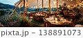 Guests are seated at wooden tables inside a wedding tent. Strings of lights hang from the top. The view shows water and city lights in the background during the evening 138891073