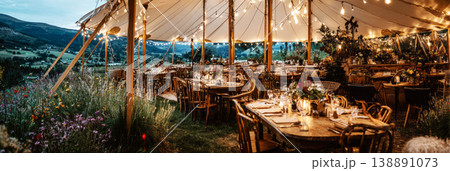 Guests are seated at wooden tables inside a wedding tent. Strings of lights hang from the top. The view shows water and city lights in the background during the evening 138891073