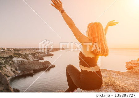 Woman sunset cliff sitting on a rocky edge with her arms raised to celebrate freedom and travel while overlooking the beautiful coastal sea Woman sunset cliff sitting on a rocky edge with her arms raised to celebrate freedom and travel while overlooking the beautiful coastal sea 138895727