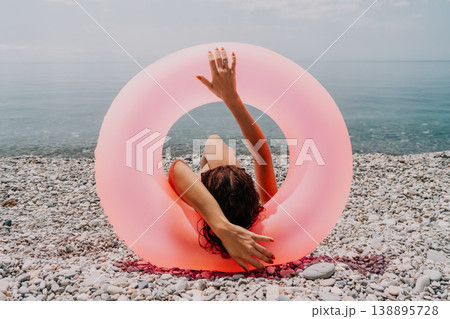 Woman beach float Sitting inside a pink inflatable ring on a pebble shore overlooking the calm blue sea during summer vacation Woman beach float Sitting inside a pink inflatable ring on a pebble shore overlooking the calm blue sea during summer vacation 138895728