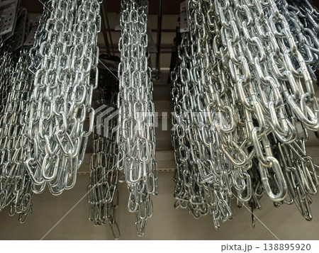 Multiple heavy duty galvanized steel chains are hanging vertically in a warehouse space. The chains form a pattern with shiny metallic links in bright light. Multiple heavy duty galvanized steel chains are hanging vertically in a warehouse space. The chains form a pattern with shiny metallic links in bright light. 138895920
